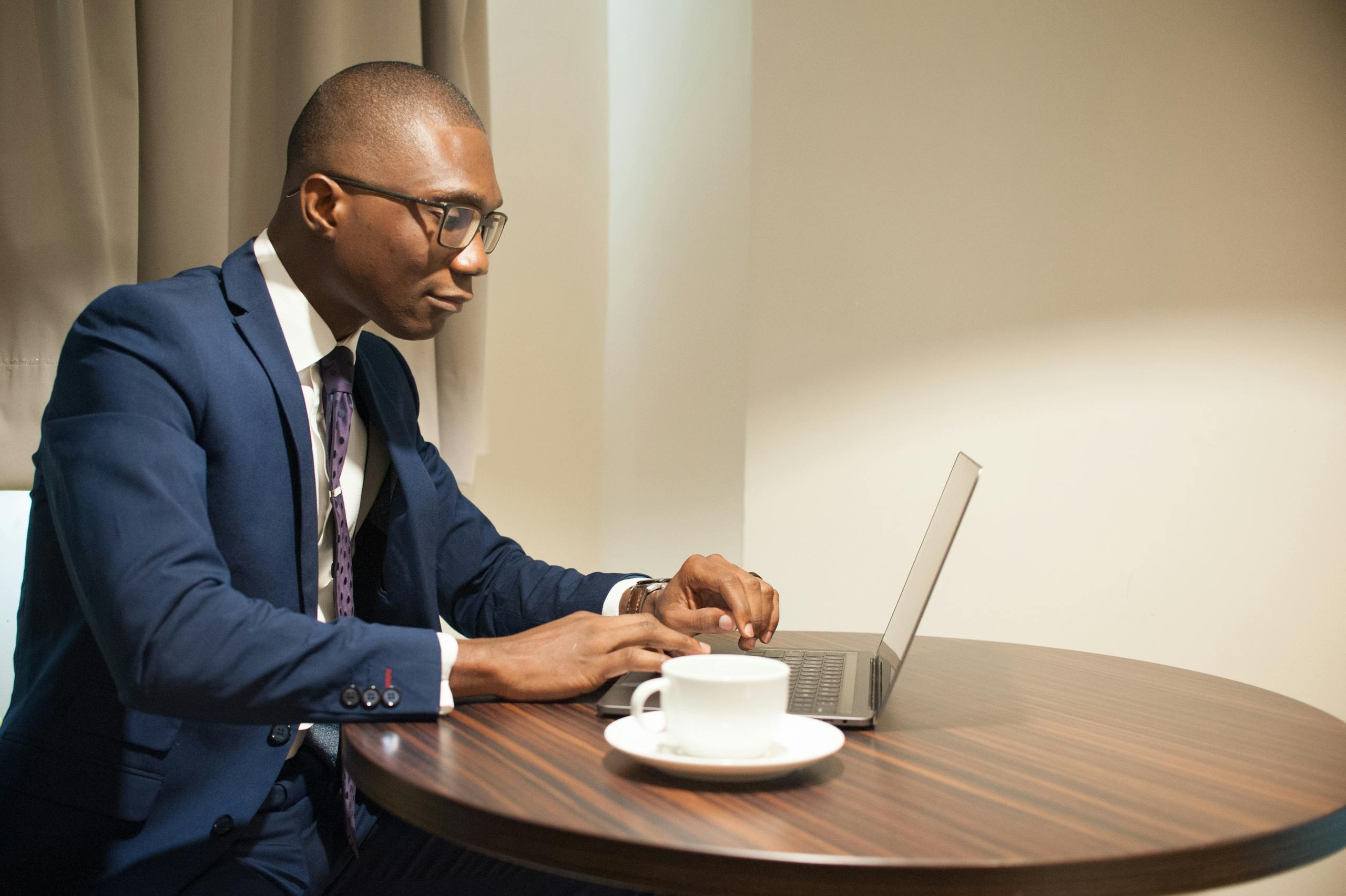 African American businessman in suit working on laptop at a round table, with a cup of coffee, indoors.